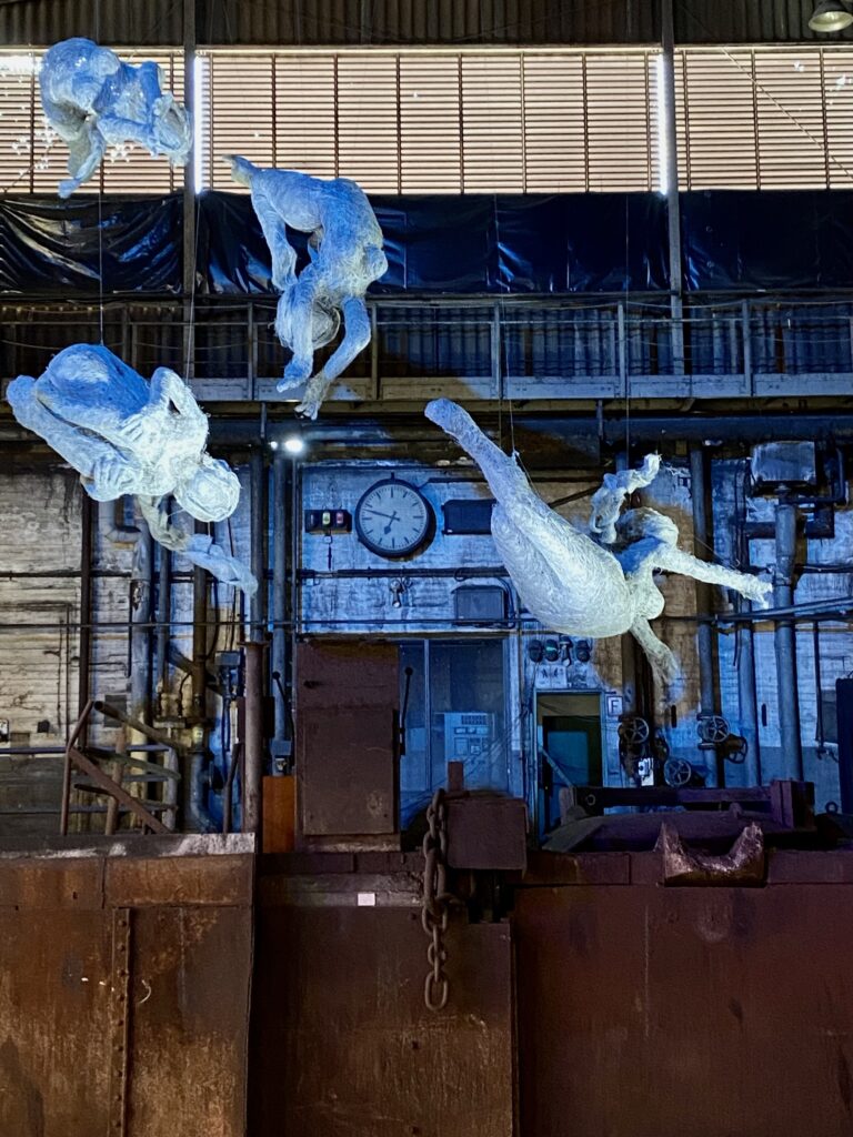 Blau leuchtende Drahtfiguren schweben vor der Wand einer Fabrikhalle